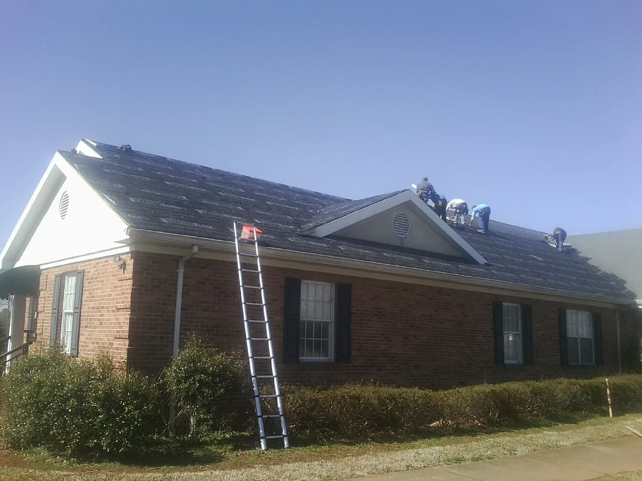 Crew tearing shingles off a brick ranch roof with a ladder set — Metal Roof Repair work by our crew in North Pitcher