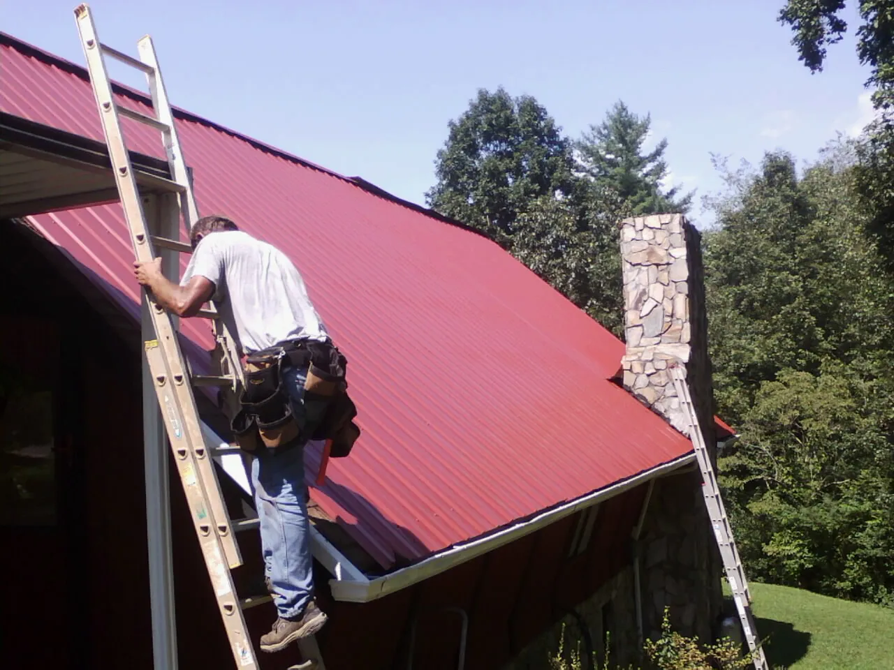Roofer on a ladder beside a red metal roof and stone chimney — Soffit & Fascia Repair by Roofing Brothers in North Pitcher
